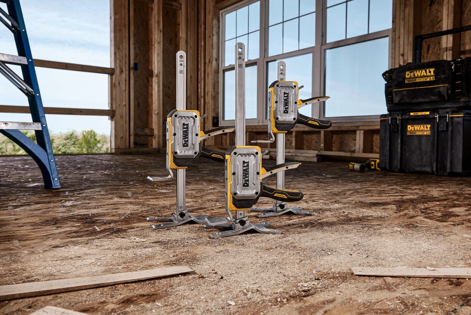Three DEWALT DWHT83550 clamps are arranged on the floor of a construction site with unfinished wooden walls and large windows. DEWALT storage boxes and a blue ladder are visible in the background.