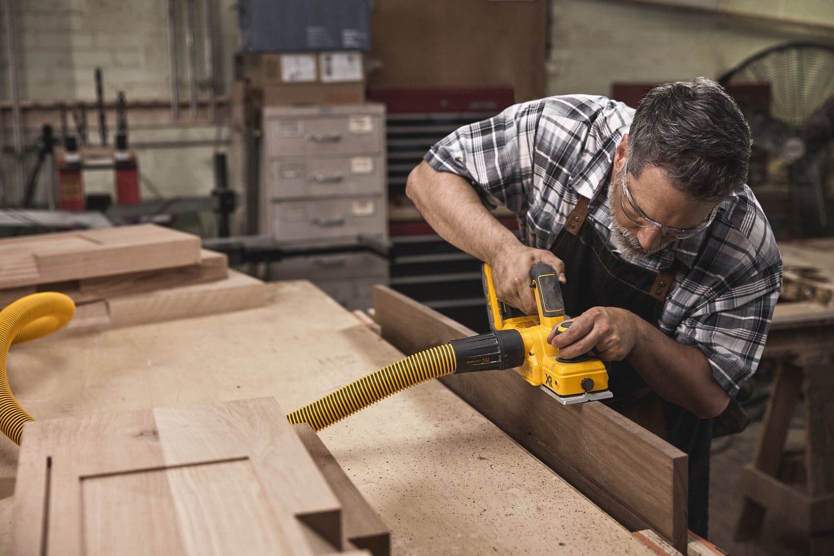 Una persona usa una cepilladora inalámbrica DEWALT (modelo DCP580B) para alisar una tabla de madera en un taller. La herramienta está conectada a una manguera amarilla para polvo, y hay varios materiales de carpintería y gabinetes en el fondo.