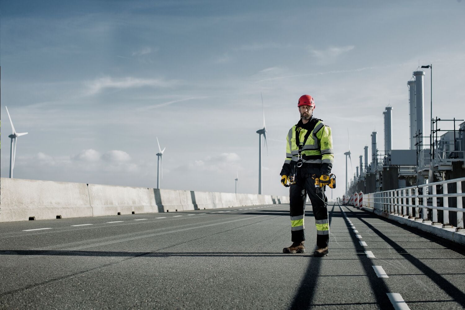 Un trabajador con ropa protectora de alta visibilidad y casco rojo se encuentra en una carretera industrial, sosteniendo herramientas DEWALT. En el fondo se pueden ver turbinas eólicas y estructuras de fábrica en un día despejado.