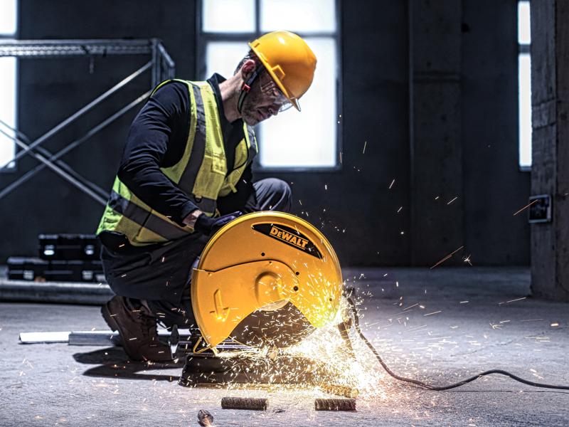 Un trabajador con casco amarillo y chaleco de seguridad utiliza una máquina de corte amarilla DEWALT D28730 para cortar tubos de metal en un entorno industrial, haciendo que salten chispas.