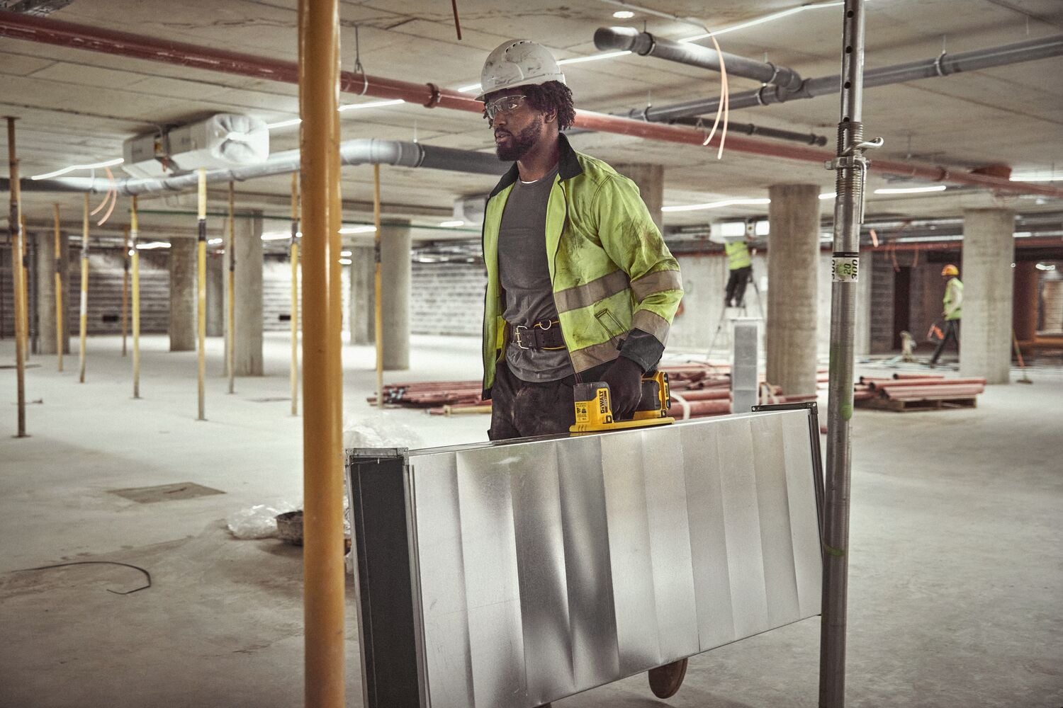 Un trabajador de la construcción con casco de seguridad y chaqueta reflectante lleva un gran panel metálico utilizando una herramienta de mango amarillo en un edificio en construcción. El rostro del trabajador está desenfocado. El entorno incluye pilares de concreto y tuberías expuestas.