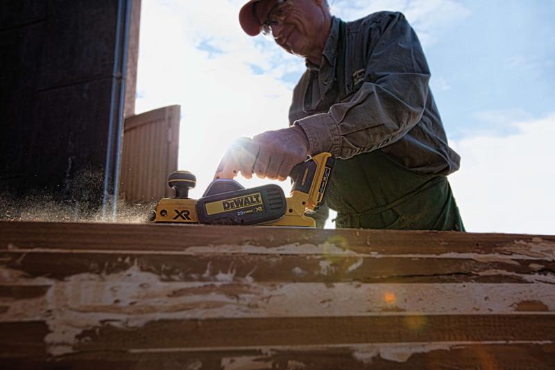 Una persona alisando una superficie de madera con una cepilladora inalámbrica DEWALT XR 20V al aire libre, mientras sale polvo durante el funcionamiento de la herramienta.