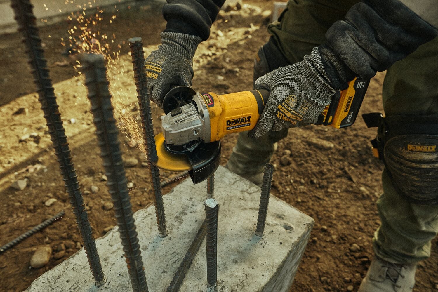 Una persona con guantes utiliza una amoladora angular inalámbrica amarilla DEWALT para cortar varillas de acero que sobresalen de un bloque de concreto, mientras se ven chispas durante el proceso de corte.