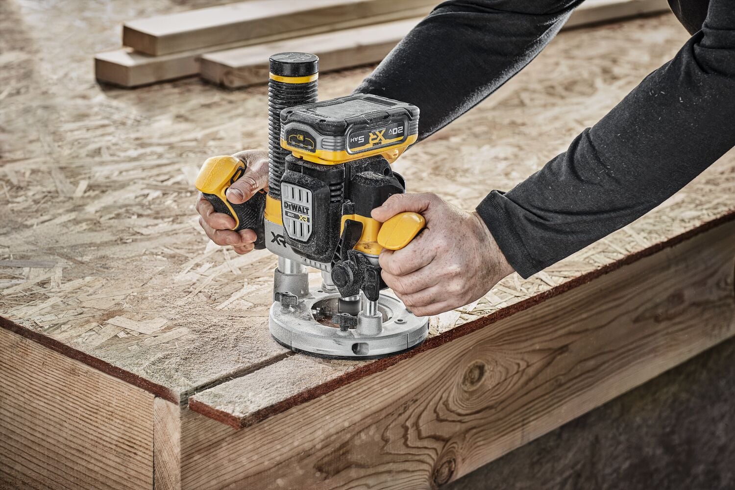 A person using a DEWALT cordless router (SKU: DCW620B) to shape the edge of a wooden board on a construction site.