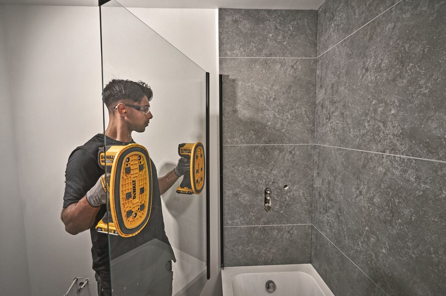 Una persona utiliza una herramienta de succión amarilla DEWALT DCE592B mientras instala o ajusta un panel de vidrio en un baño con paredes de baldosas grises.