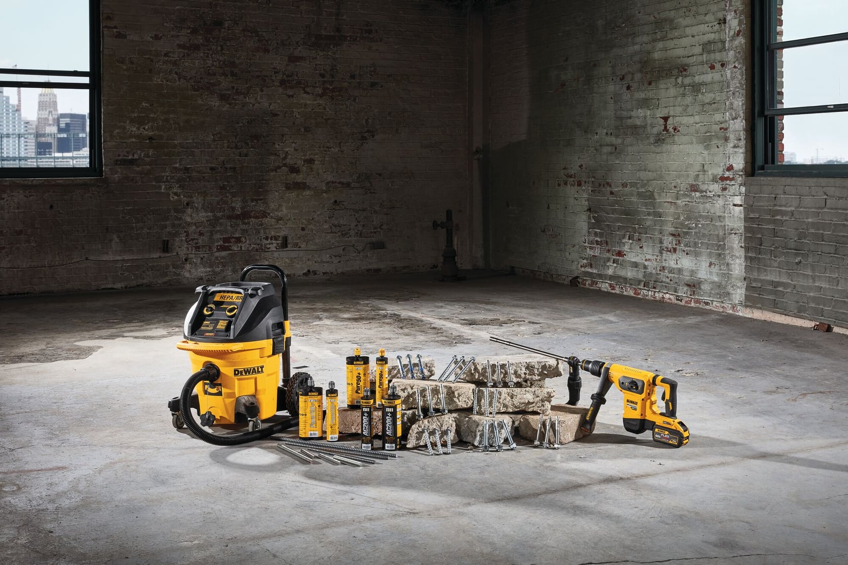 Un conjunto de herramientas y accesorios de la marca DEWALT, incluyendo una aspiradora de seco/húmedo, una herramienta eléctrica, brocas y botellas de lubricante, dispuestas sobre un suelo de hormigón en un espacio industrial con paredes de ladrillo y ventanas grandes.