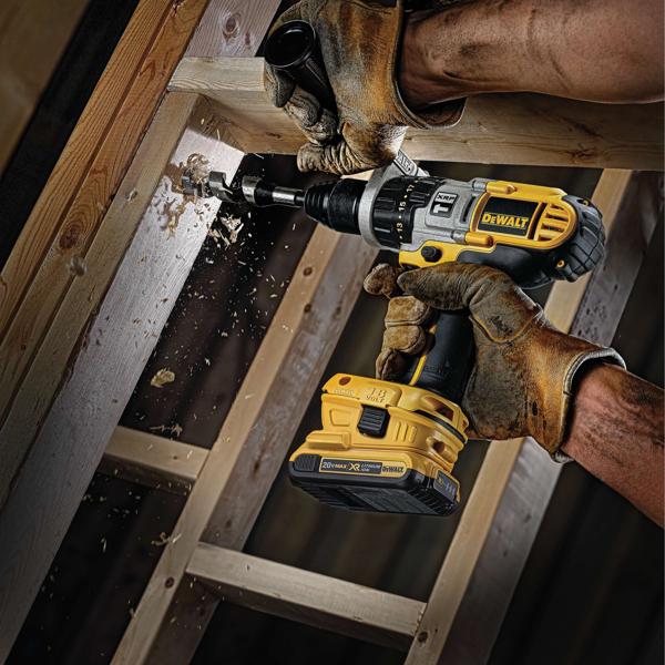 Una persona con guantes de trabajo utiliza un taladro inalámbrico DEWALT para perforar un agujero en una viga de madera, mientras vuelan astillas de madera durante el funcionamiento del taladro.
