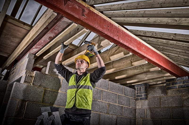 Un trabajador de la construcción con casco de seguridad amarillo y chaleco reflectante verde utiliza un taladro inalámbrico DEWALT para fijar vigas de madera a un soporte de acero rojo dentro de un edificio en construcción. Las paredes están hechas de bloques de hormigón y se pueden ver vigas de madera arriba.