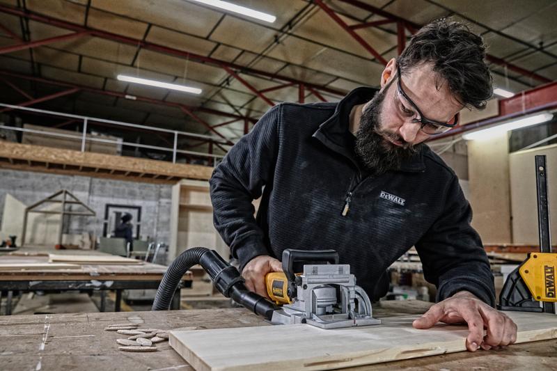 Una persona utiliza una herramienta de ensamblaje DEWALT DW682K para trabajar una pieza de madera en un taller. El rostro de la persona está difuminado por privacidad. El espacio de trabajo incluye mesas, piezas de madera y equipos de taller visibles.