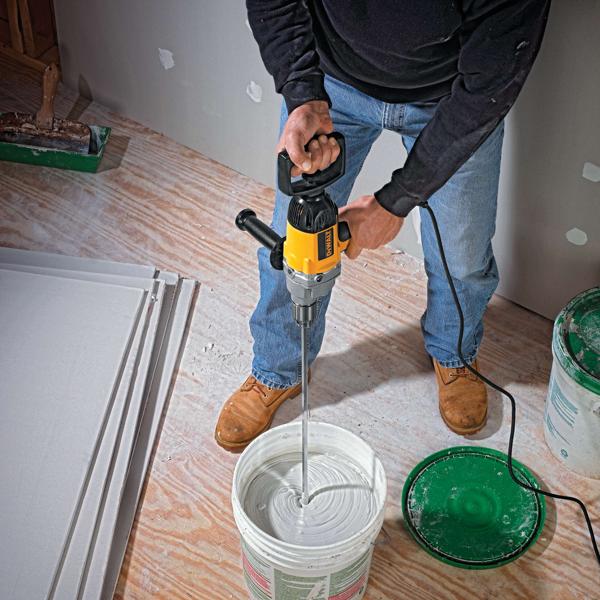 Una persona utiliza una mezcladora eléctrica DEWALT para mezclar un compuesto blanco en un balde grande en una obra. La persona está de pie sobre un piso de madera, usando botas de trabajo, jeans y un suéter negro. Hay láminas de panel de yeso y materiales de construcción cerca.