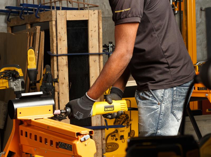 Una persona con guantes utiliza una herramienta esmeriladora recta DEWALT DWE4887N sobre una pieza de trabajo en un taller. Se pueden ver varias herramientas y equipos de taller amarillos DEWALT en el fondo.