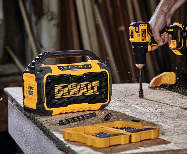 Un altavoz Bluetooth portátil DEWALT está colocado sobre un banco de trabajo junto a una caja de herramientas amarilla con brocas. Al fondo, una persona utiliza un taladro DEWALT para perforar la superficie de madera, generando virutas. La escena ocurre en un taller.