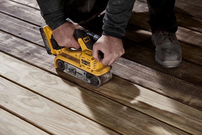 Una persona está usando una lijadora de banda inalámbrica amarilla DEWALT para lijar tablas de madera de una tarima.