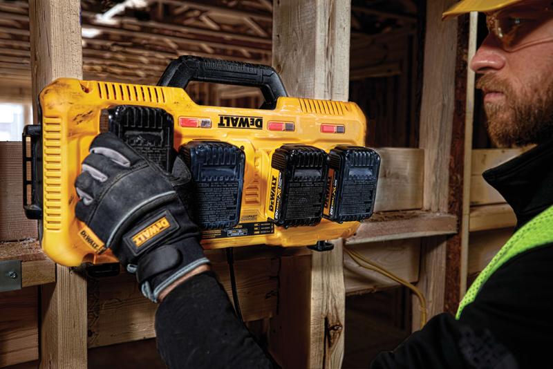 Un cargador de baterías multiport amarillo Dewalt DCB104 montado en una viga de madera en un sitio de construcción. Una persona con guantes con la marca Dewalt está manejando el cargador, y hay cuatro baterías Dewalt conectadas.