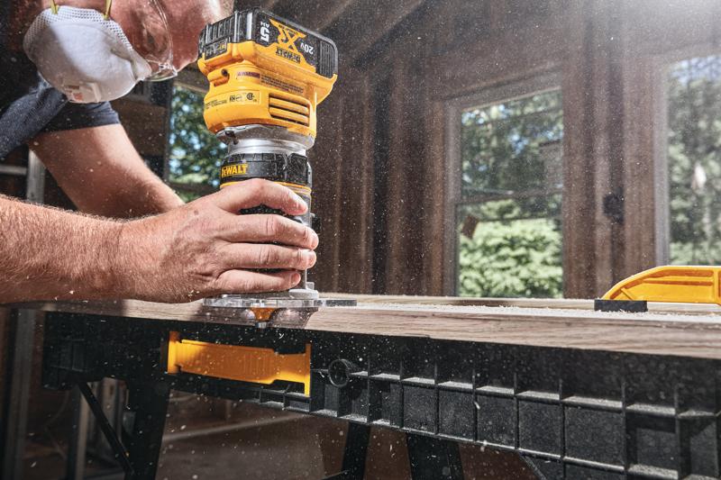 Una persona con mascarilla antipolvo utiliza una fresadora inalámbrica amarilla DEWALT sobre una pieza de madera, generando polvo en un taller.