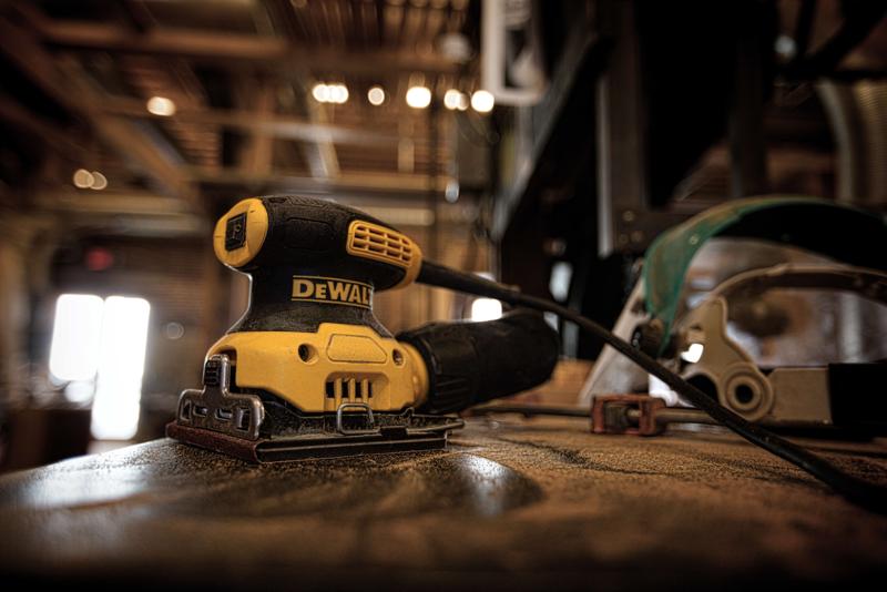 Una lijadora eléctrica DEWALT DWE6411K está sobre una mesa de trabajo polvorienta en un taller, con un cable conectado y equipo de seguridad visible en el fondo.