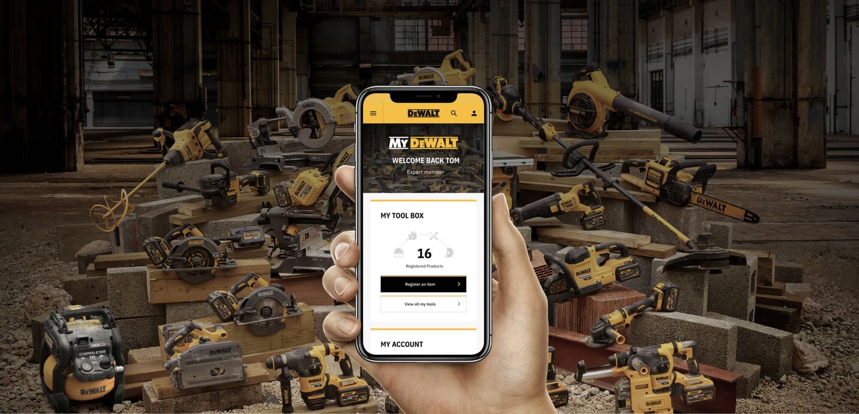 Una mano sostiene un teléfono inteligente que muestra la página de cuenta My DEWALT, con productos registrados. Al fondo, hay una gran variedad de herramientas eléctricas DEWALT organizadas en un taller industrial.