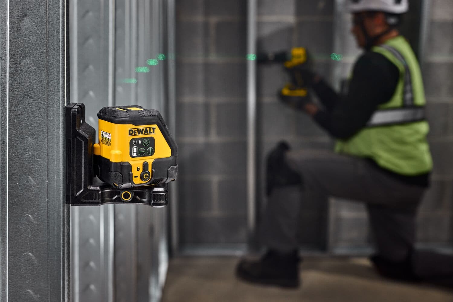 Una herramienta de nivel láser DEWALT montada en un marco de pared de metal, proyectando una línea láser verde a través de la habitación. Al fondo, una persona con casco de seguridad y chaleco reflectante trabaja con otra herramienta DEWALT, arrodillada cerca del marco de metal.