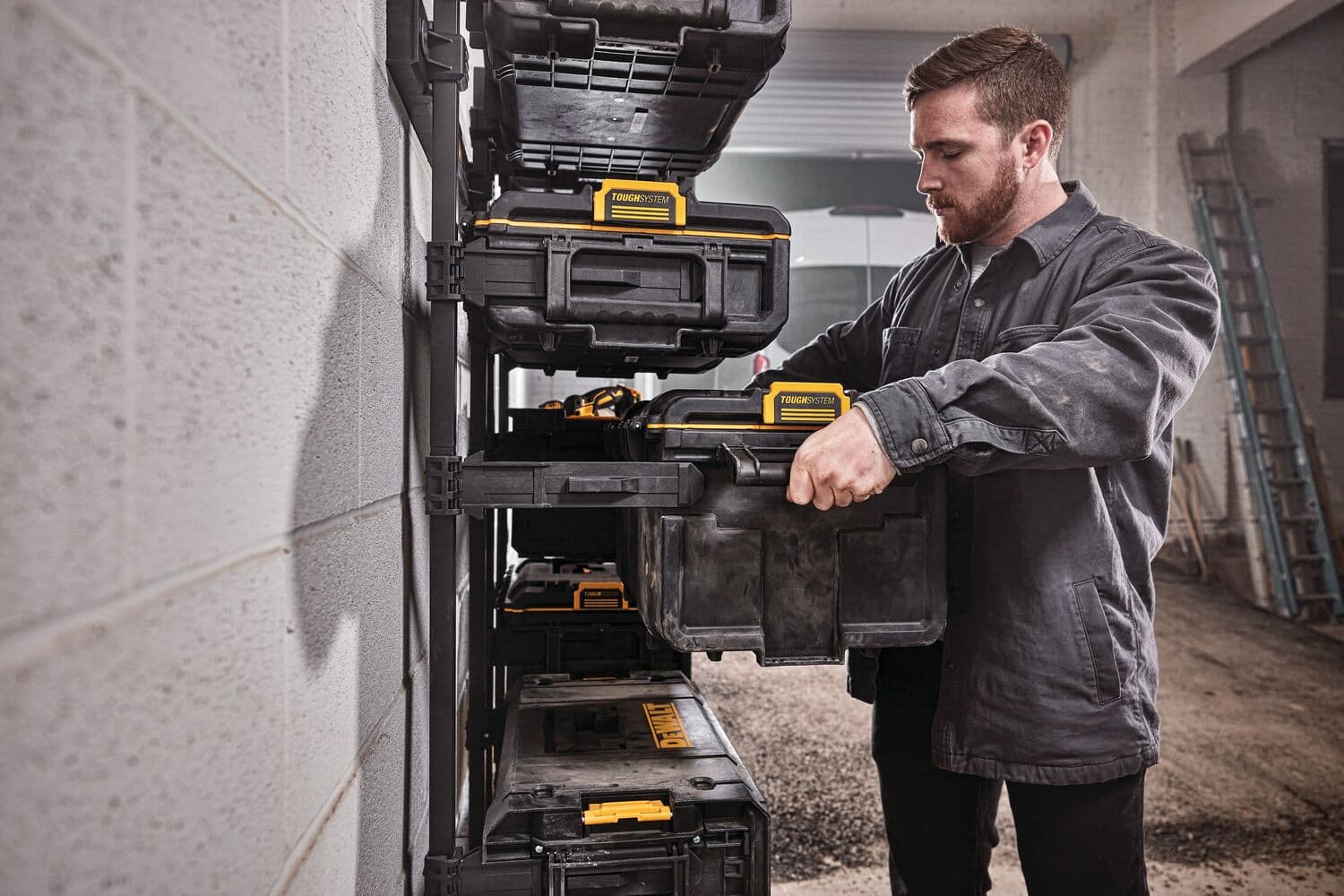 Una persona organiza cajas de almacenamiento DEWALT en un estante montado en la pared de un taller o garaje. El rostro de la persona está difuminado y lleva una chaqueta de trabajo oscura.