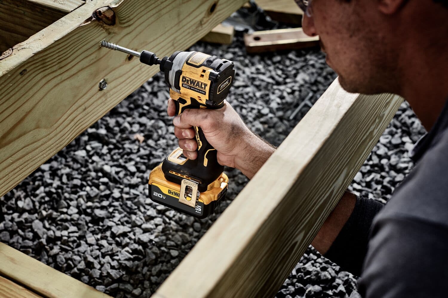 A person is using a DEWALT DCF850B cordless impact driver to drill a screw into a piece of wooden framing outdoors, with gravel on the ground.