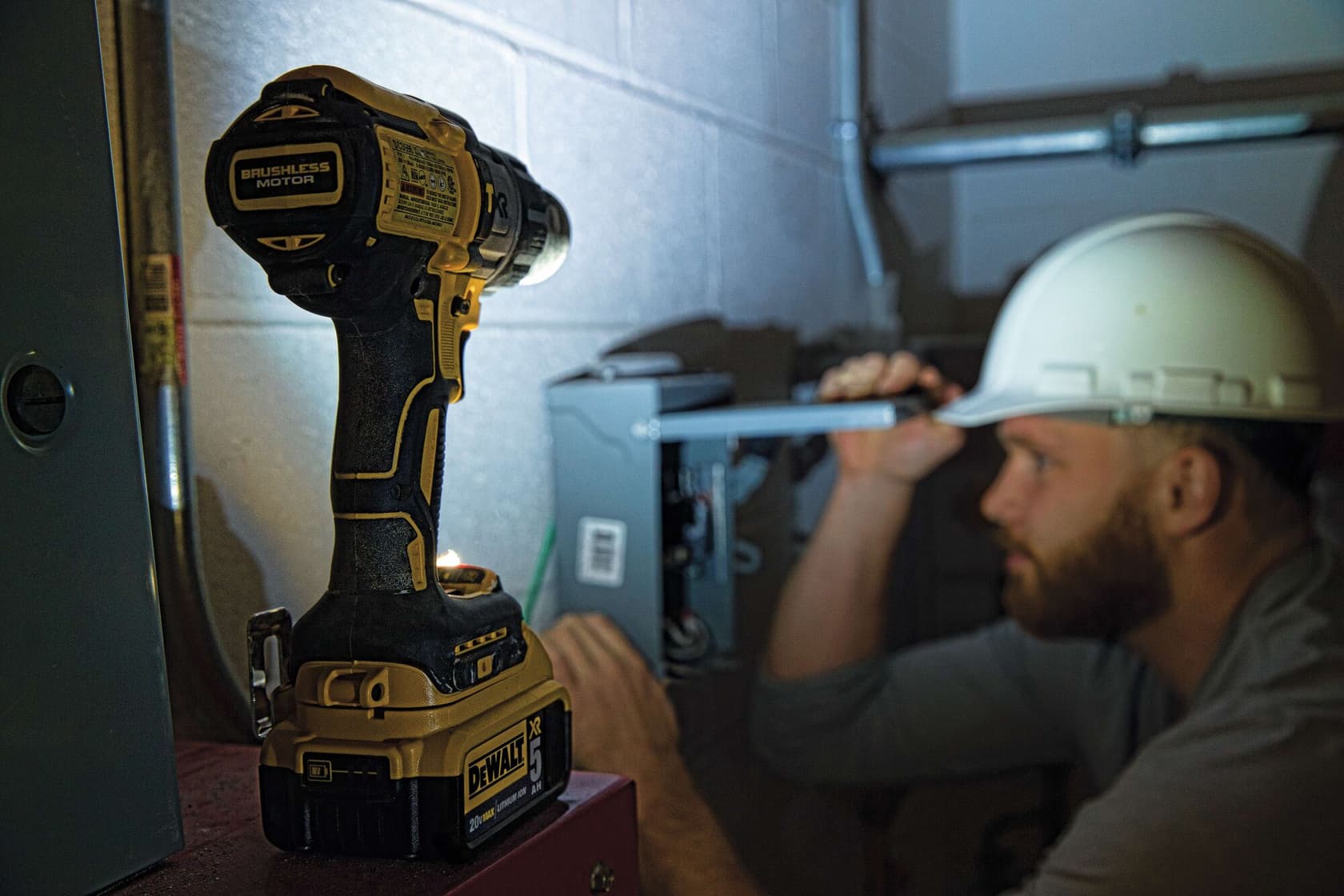 Un taladro inalámbrico DEWALT con motor sin escobillas se muestra en primer plano, colocado sobre una superficie, con una persona usando casco trabajando en equipo eléctrico en el fondo.