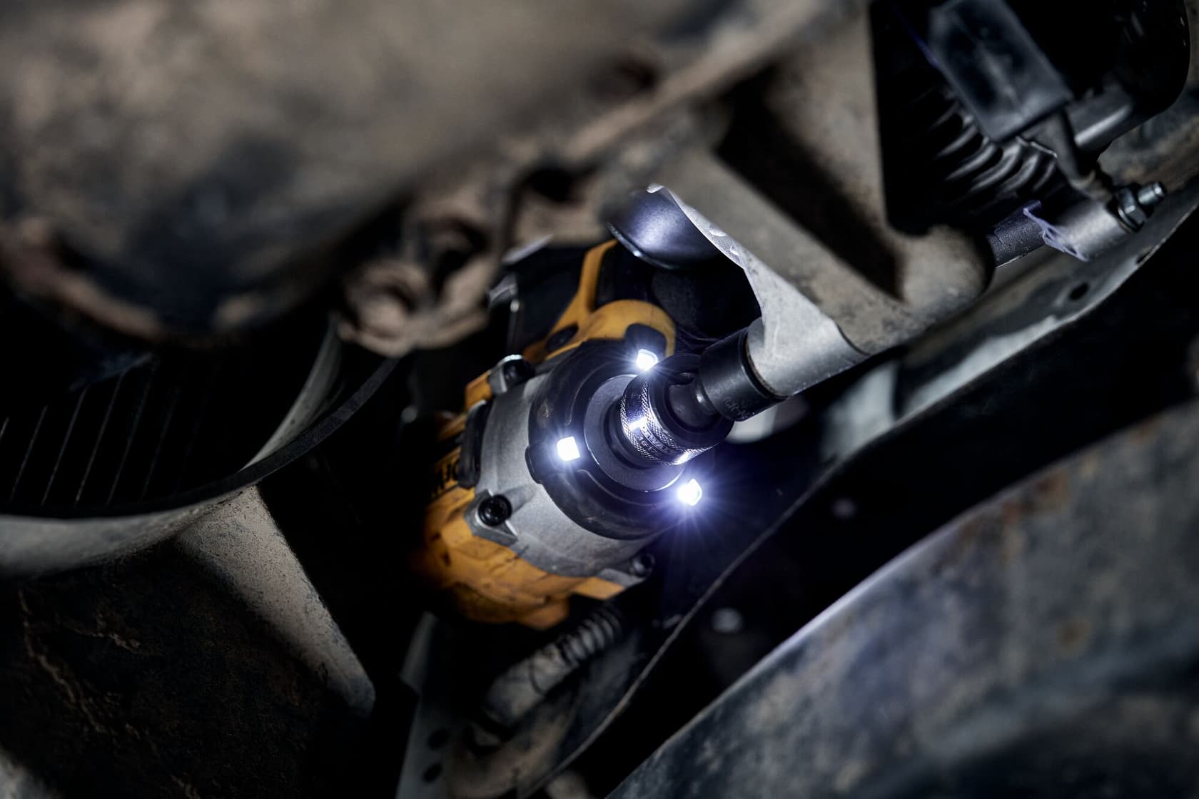 A close-up view of a DEWALT cordless impact wrench being used on a mechanical part under a vehicle. The tool features bright LED lights illuminating the work area.