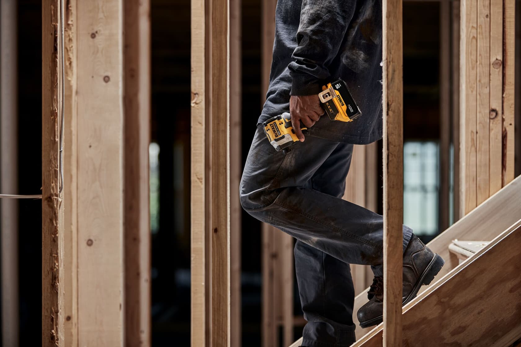 Person wearing work clothes and boots, holding a DEWALT cordless impact driver, stands on a wooden staircase inside an unfinished building framed with exposed wood studs.