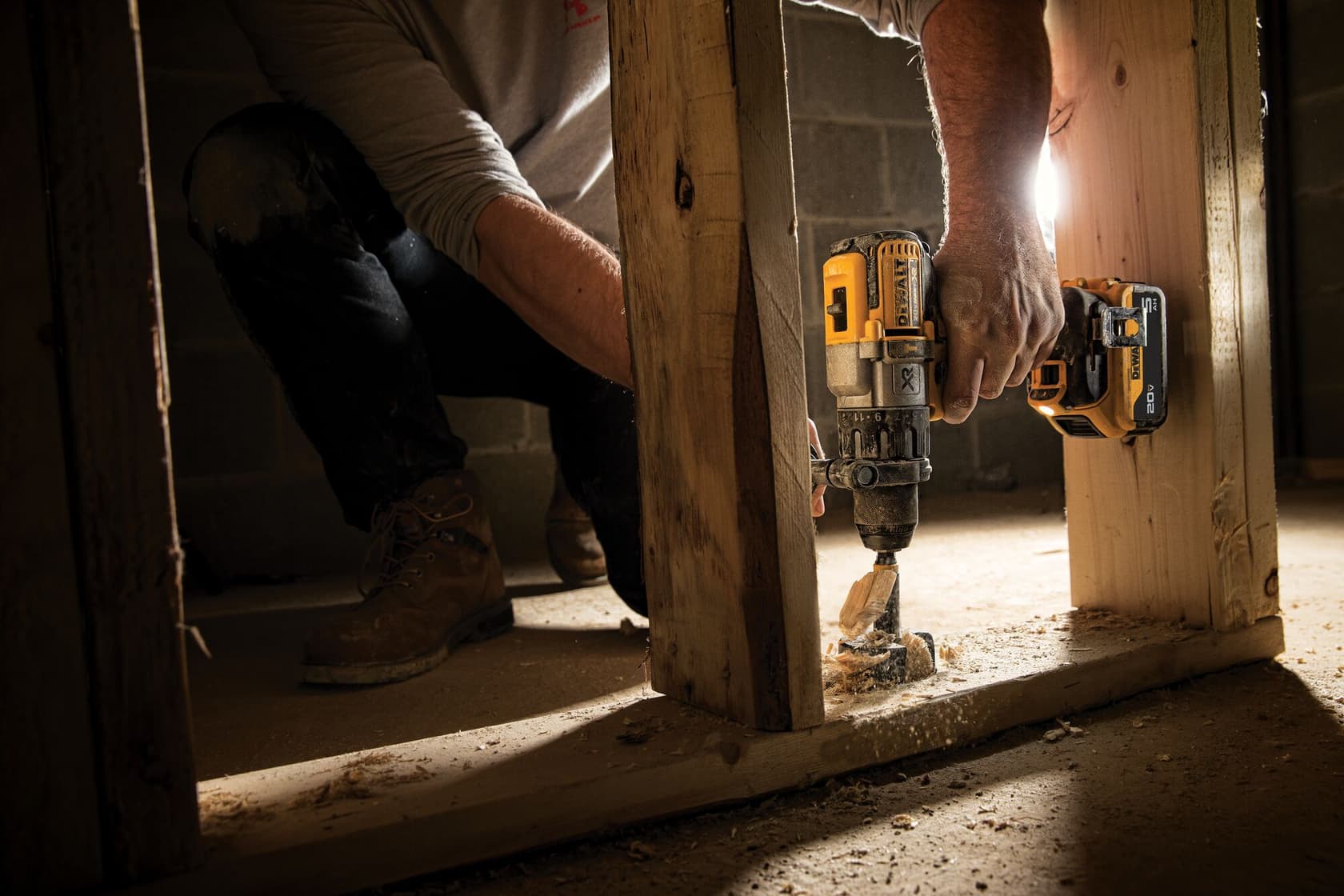 Una persona utiliza un taladro inalámbrico DEWALT para perforar una viga de madera en un sitio de construcción. El área está poco iluminada y hay virutas de madera esparcidas en el suelo.