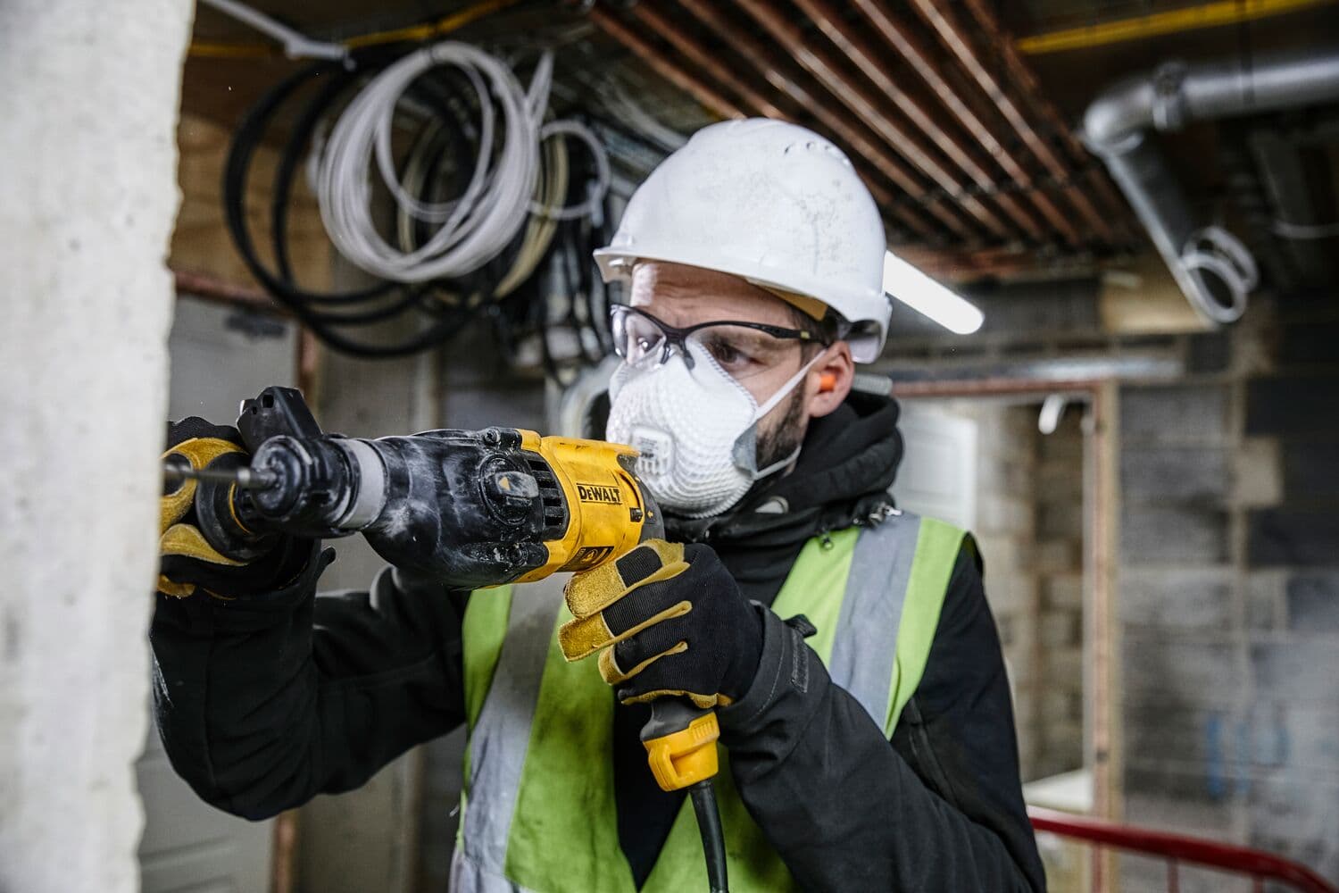 Una persona con casco de seguridad blanco, guantes y chaleco de alta visibilidad está usando un martillo rotativo DEWALT D25133K en una pared de concreto en un sitio de construcción.
