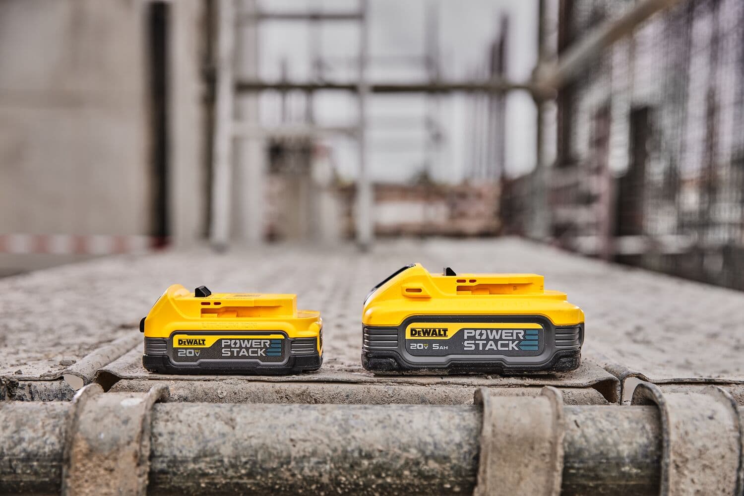 Dos baterías amarillas DEWALT Power Stack de 20V están expuestas una al lado de la otra sobre una superficie polvorienta de obra, con andamiaje industrial y cercado al fondo.