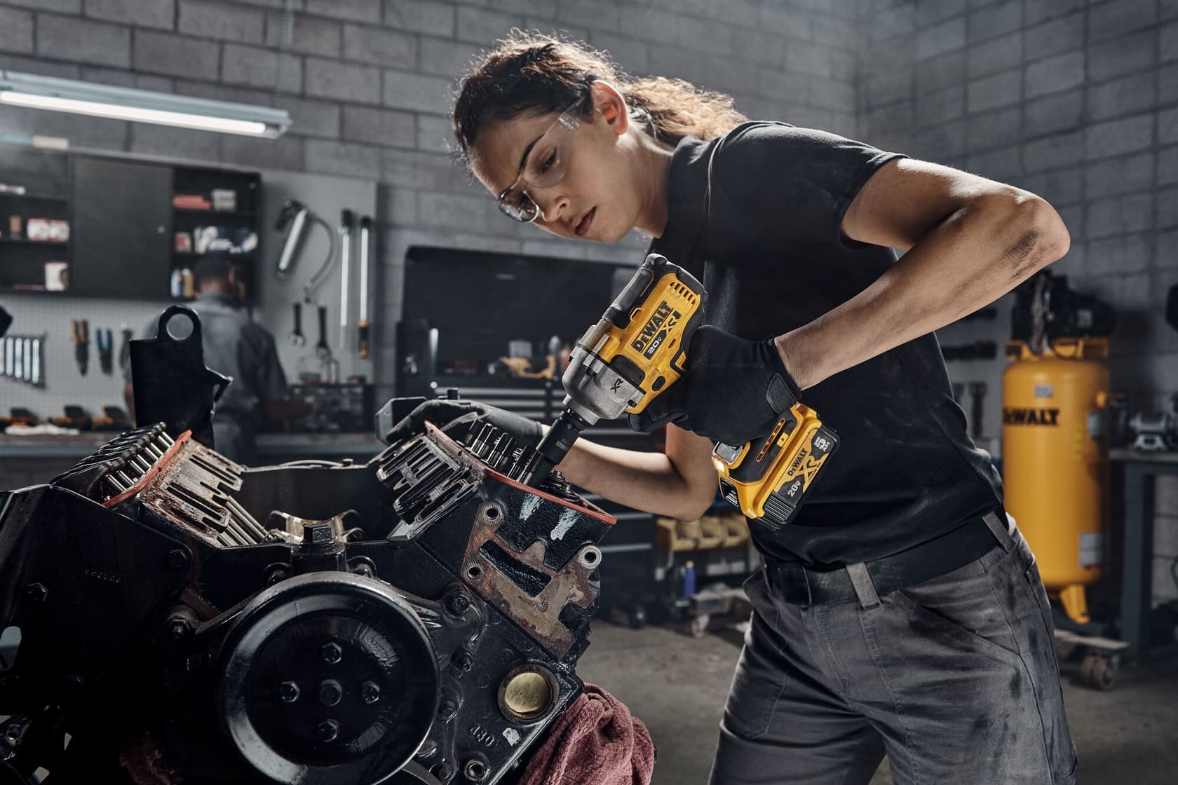 Persona usando una llave de impacto inalámbrica DEWALT (modelo DCF892B) para trabajar en un bloque de motor en un taller. El fondo muestra herramientas, estantes y un gran compresor de aire amarillo DEWALT.