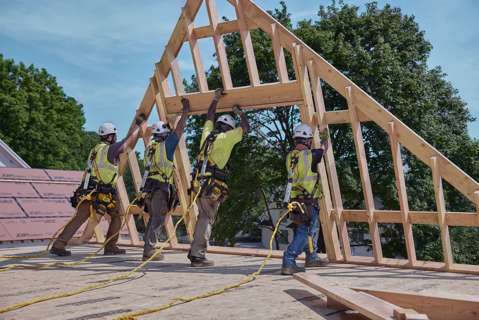 Cuatro trabajadores de la construcción con equipo de seguridad y arneses levantan y colocan una estructura de techo de madera en un sitio de construcción. Árboles y cielo despejado se ven en el fondo.