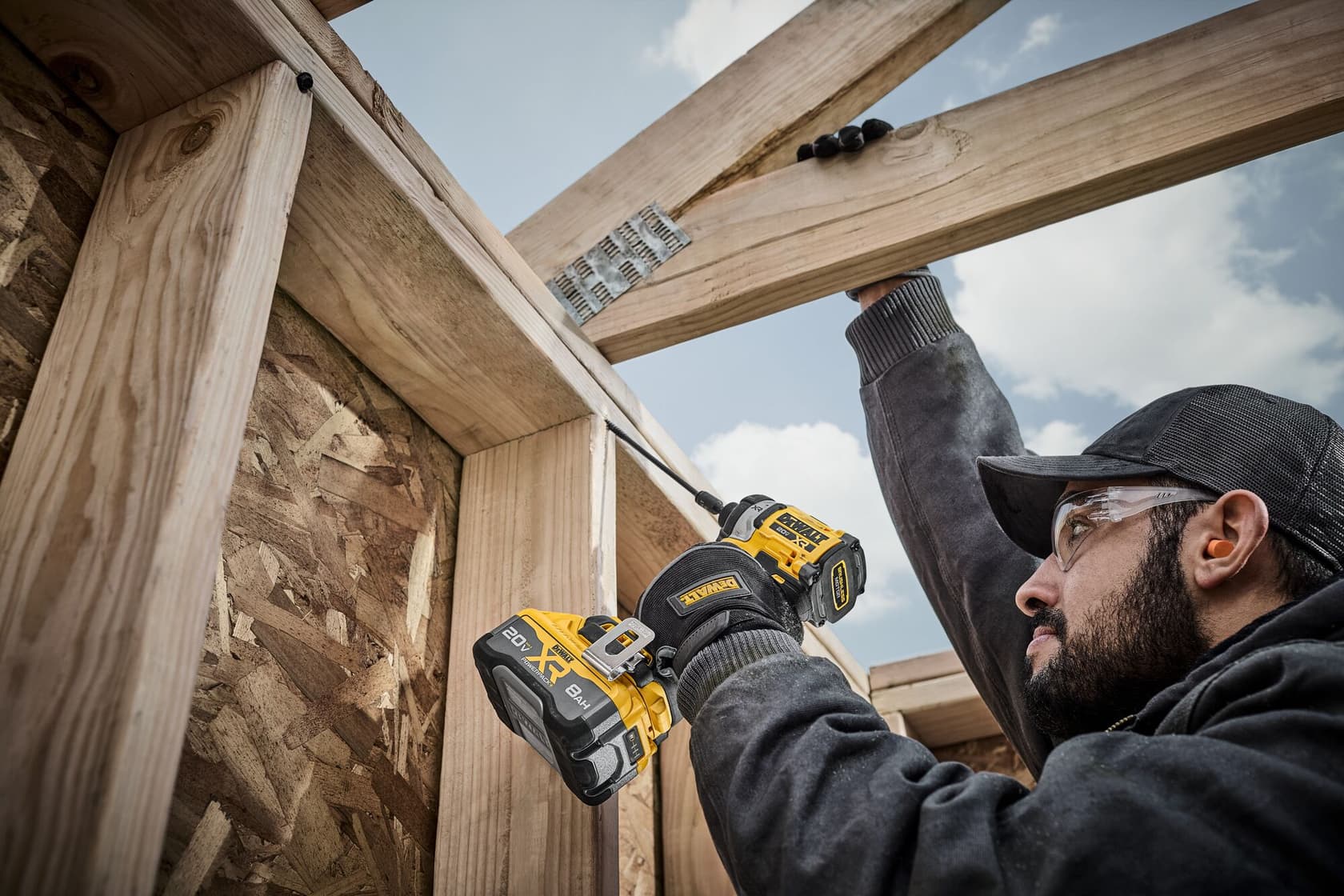 A person using a DEWALT DCF860B cordless drill to assemble wooden beams at a construction site. The individual's face is obscured, and the drill is visibly marked with the DEWALT logo.