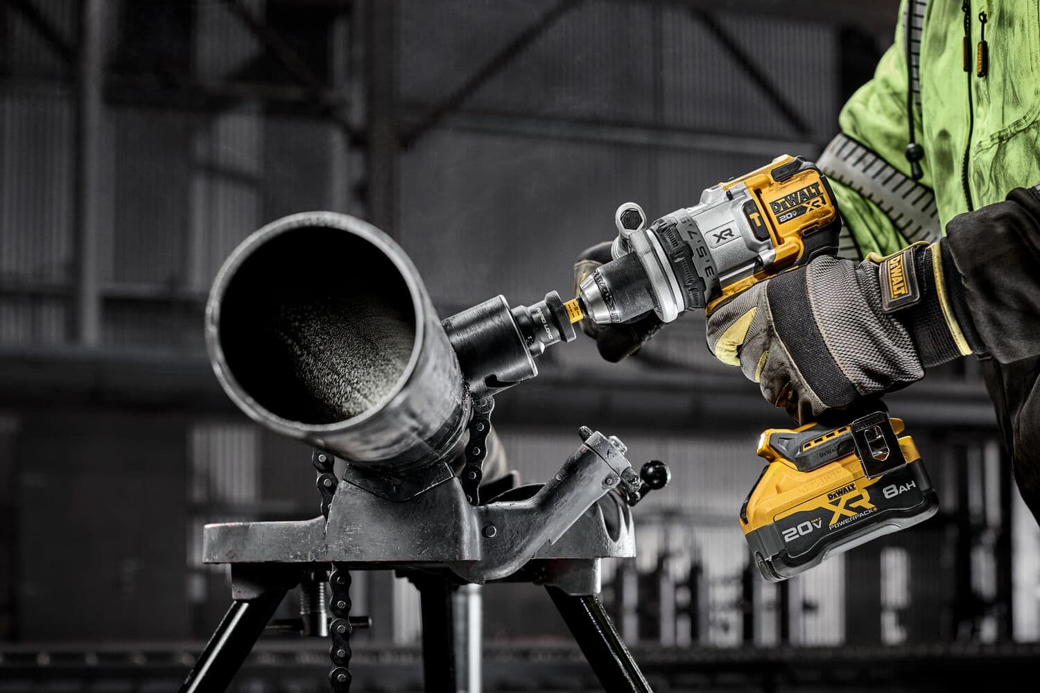 Una persona con guantes protectores y chaqueta verde está utilizando un taladro inalámbrico DEWALT para trabajar en un tubo de metal grande, que está asegurado en un soporte metálico dentro de un entorno industrial.