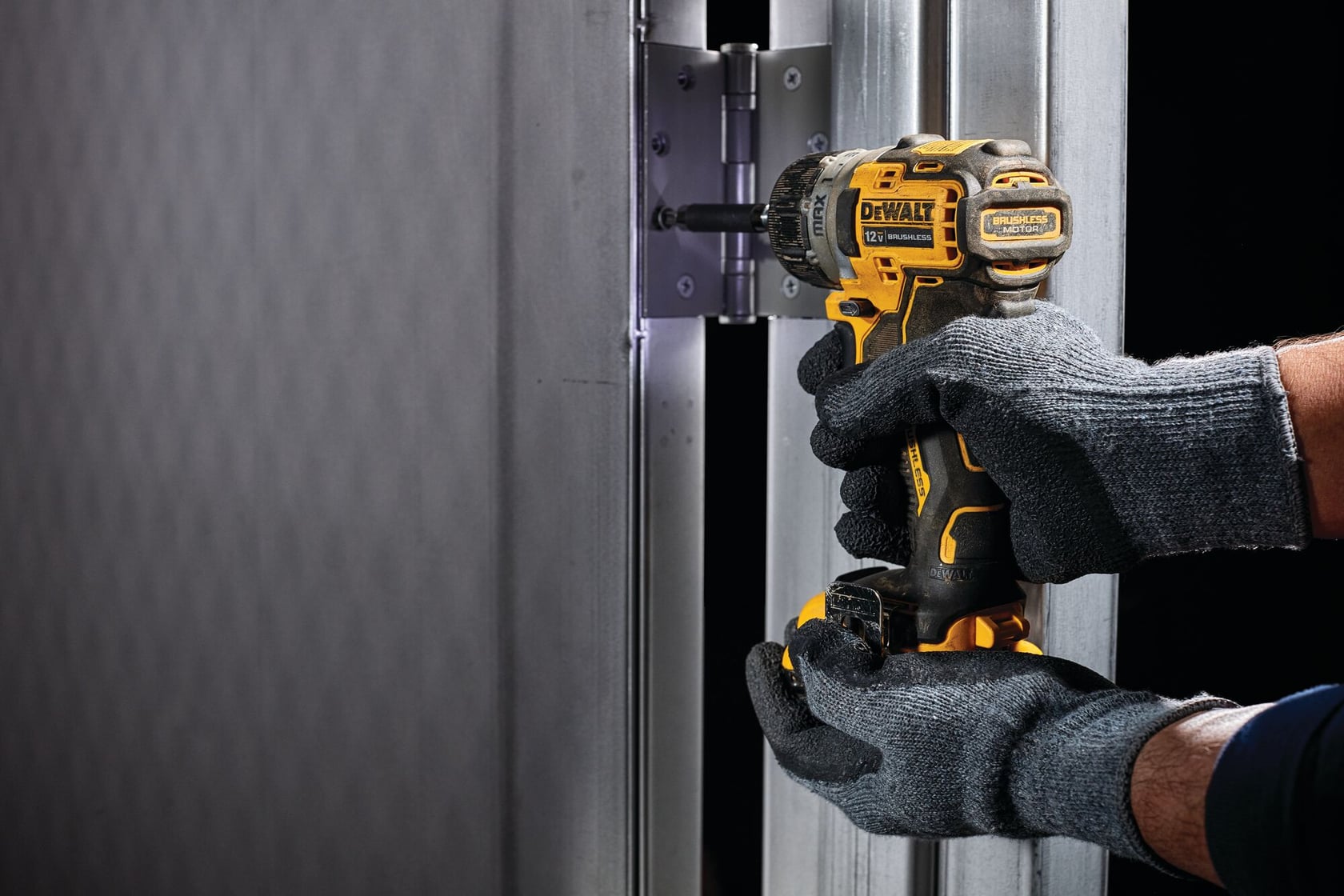 Una persona con guantes de trabajo grises utiliza un taladro inalámbrico amarillo y negro DEWALT para fijar una bisagra de metal a un marco de puerta.
