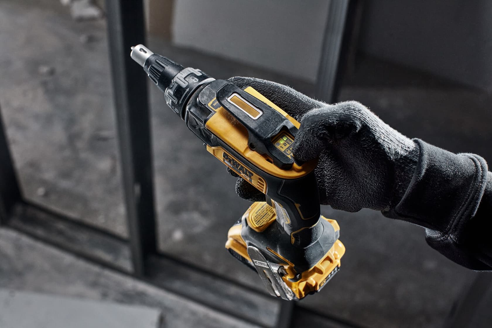 Una mano enguantada sostiene un destornillador inalámbrico DEWALT. La herramienta es de color amarillo y negro, con botones y indicadores visibles en el cuerpo, y se utiliza en un entorno interior.