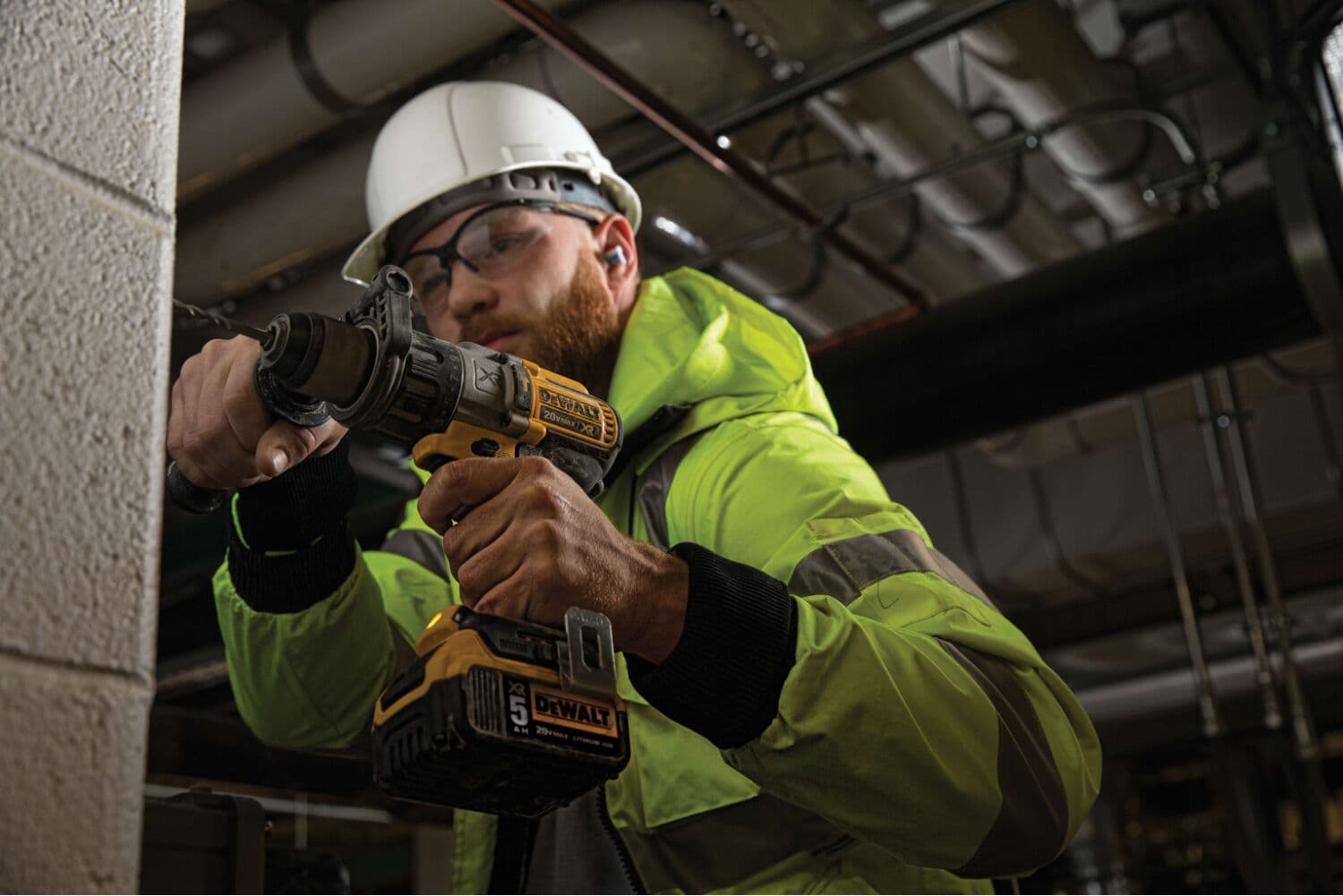 Una persona con chaqueta de seguridad verde y casco blanco utiliza un taladro inalámbrico DEWALT DCD996P2 para trabajar en una pared de concreto en un entorno industrial. El rostro está difuminado por privacidad.