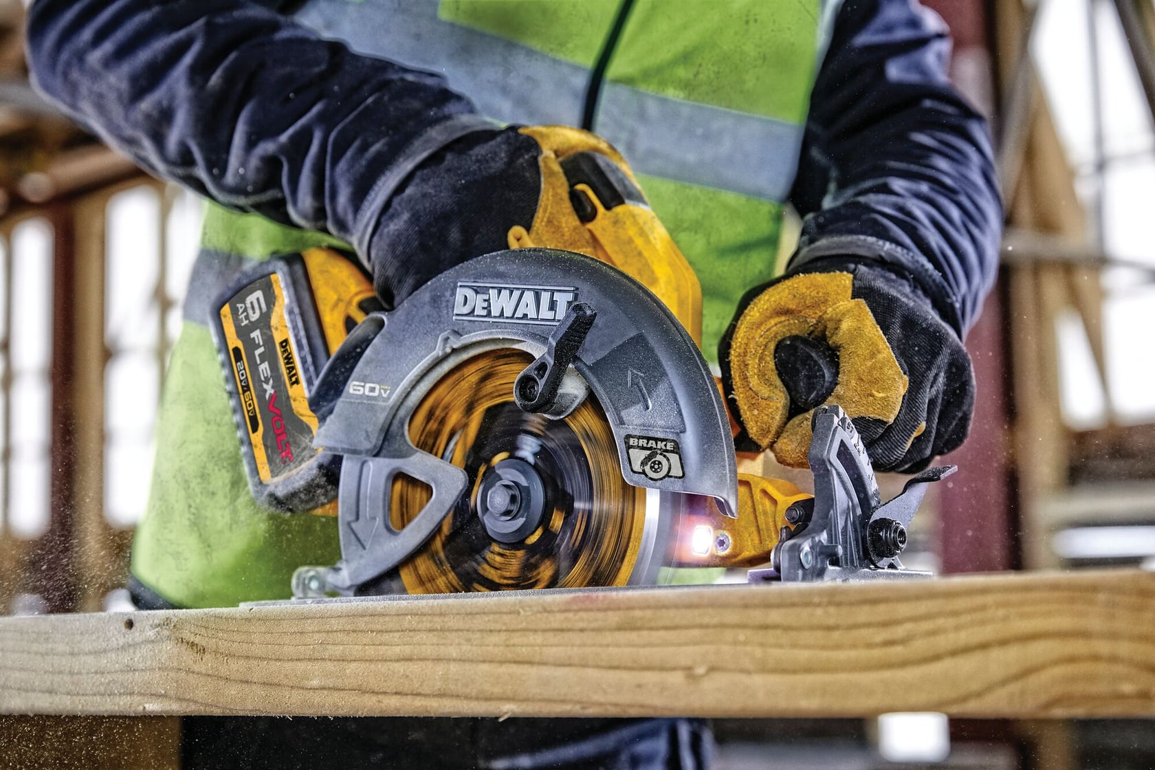 Primer plano de una persona usando una sierra circular DEWALT para cortar un trozo de madera. La persona lleva guantes de trabajo amarillos y un chaleco de seguridad brillante, con un sitio de construcción visible en el fondo.