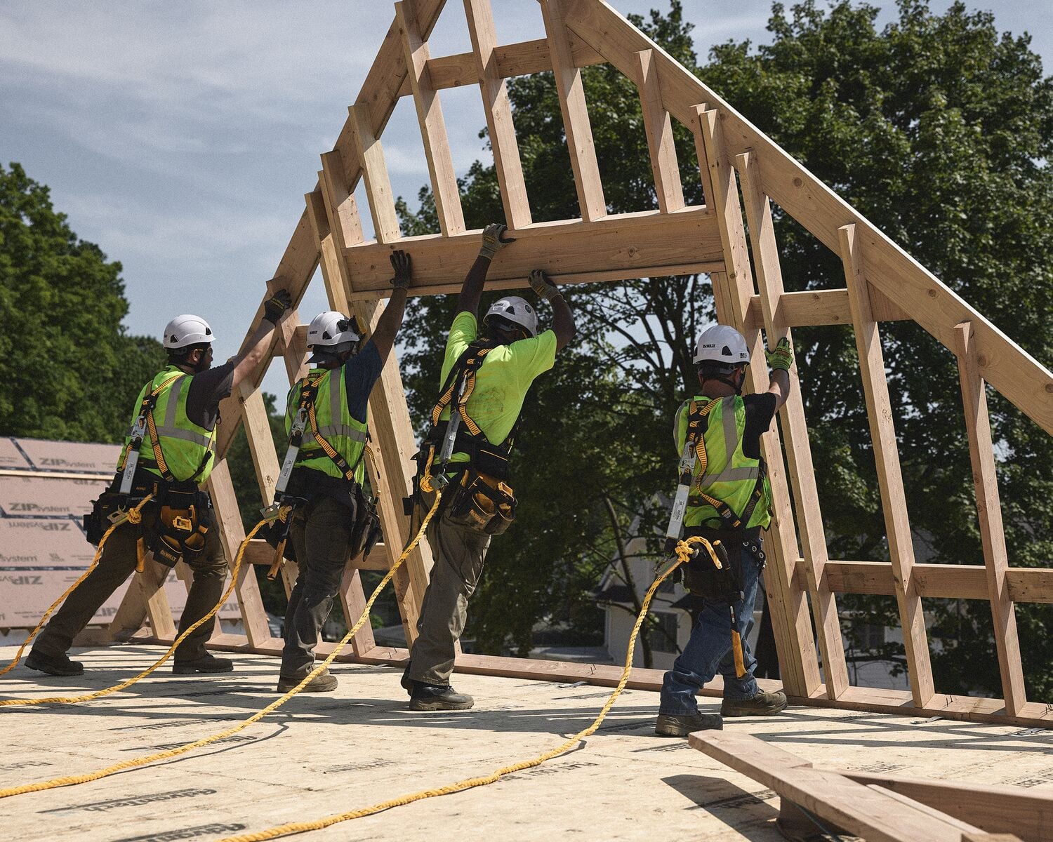 Cuatro trabajadores de la construcción con arneses de seguridad, cascos y chalecos de alta visibilidad están levantando una estructura de madera para el techo en un sitio de construcción. En el fondo se pueden ver árboles y materiales de construcción.