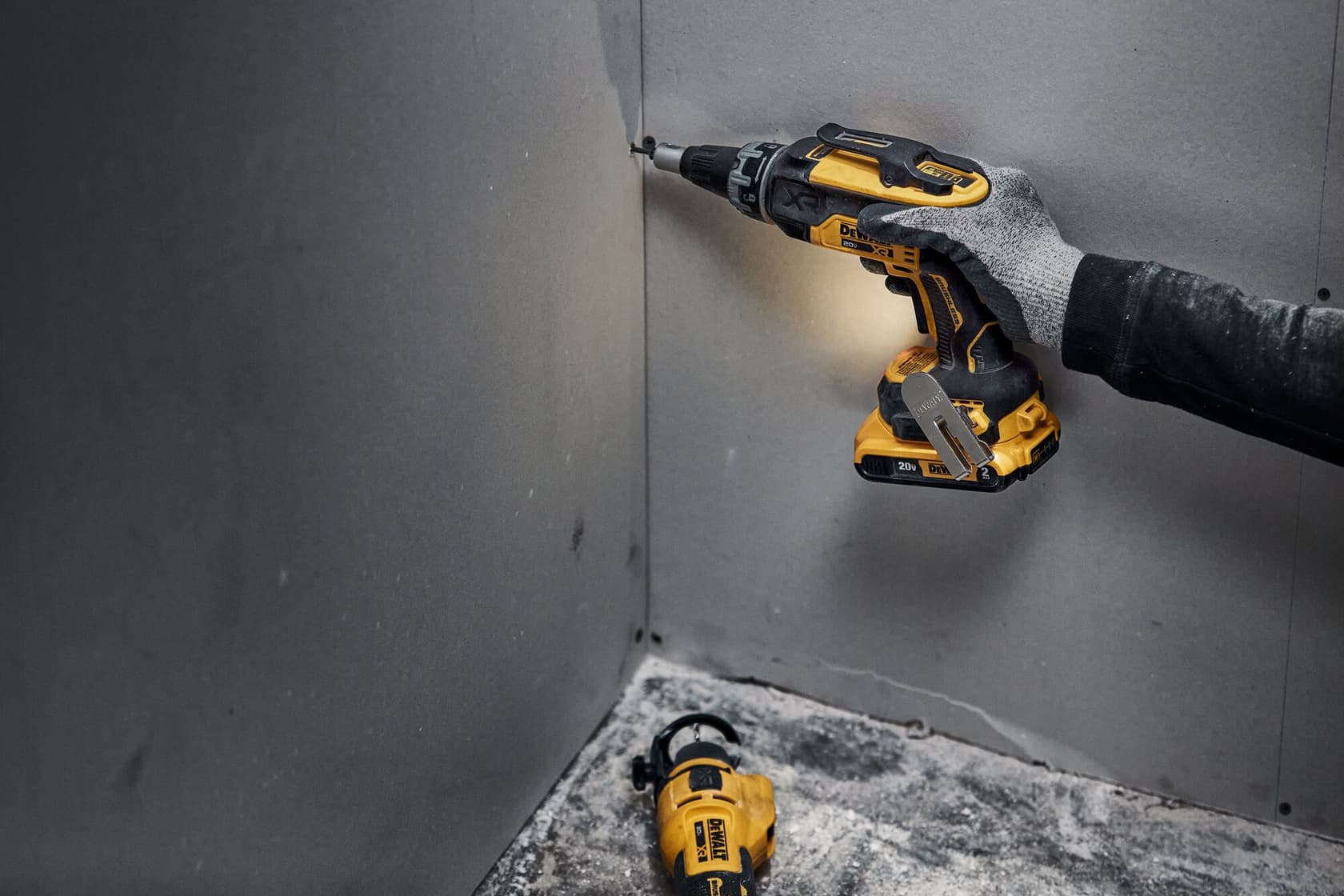 Una persona con guante utiliza un destornillador inalámbrico amarillo y negro DEWALT para atornillar en una esquina de panel de yeso. Otra herramienta amarilla DEWALT está en el suelo polvoriento cerca.