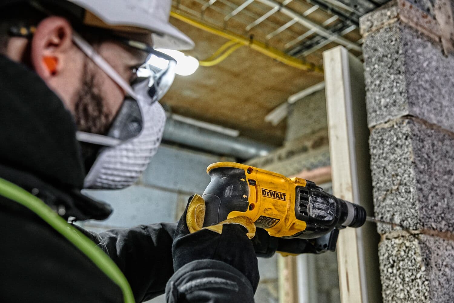 Una persona con equipo de seguridad utiliza un taladro percutor rotativo amarillo DEWALT D25133K para trabajar en una pared de concreto en un entorno de construcción.