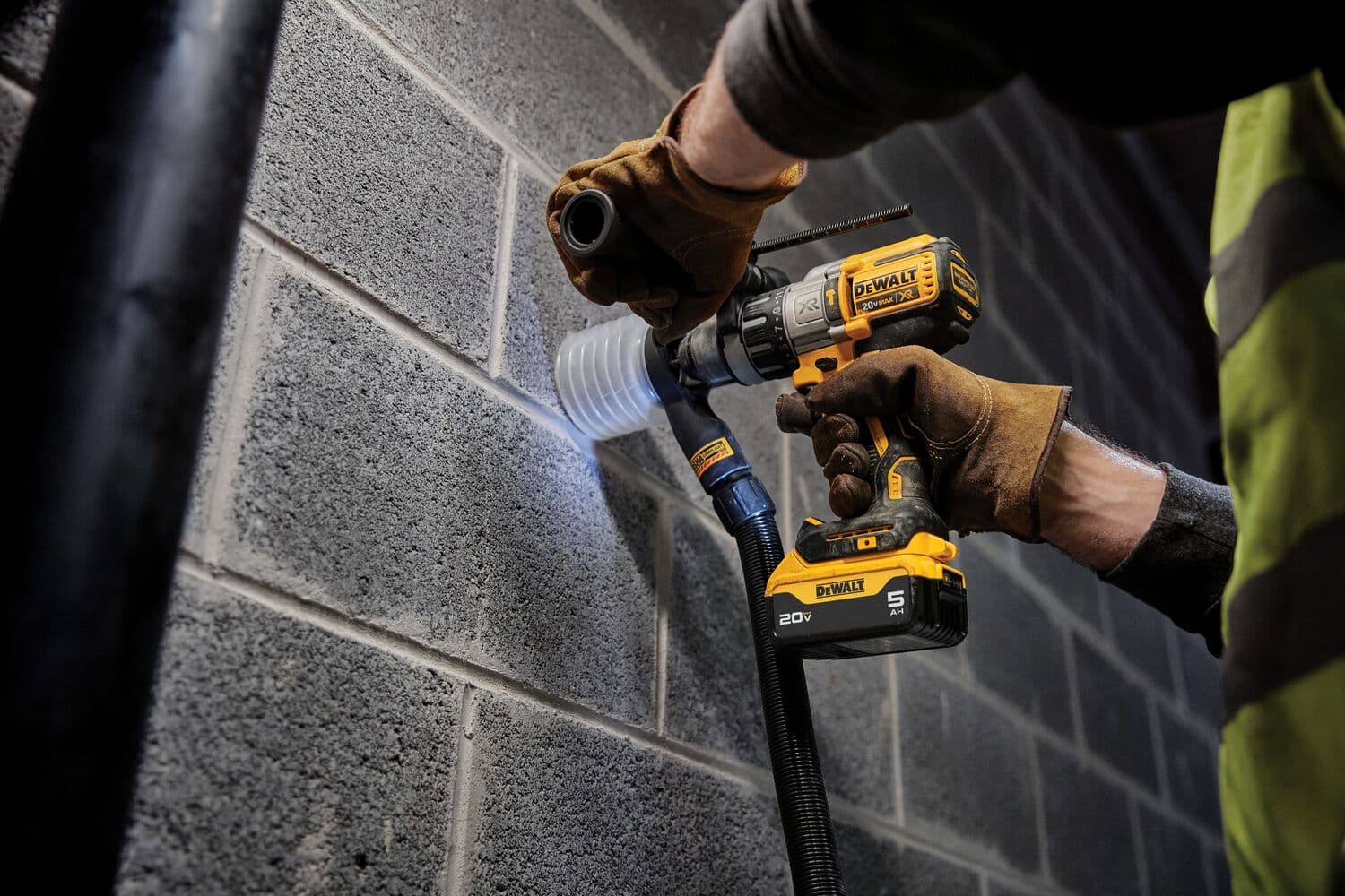 Una persona con guantes de trabajo usa un taladro inalámbrico DEWALT para hacer un agujero en una pared de bloques de concreto, con un accesorio de extracción de polvo conectado al taladro.