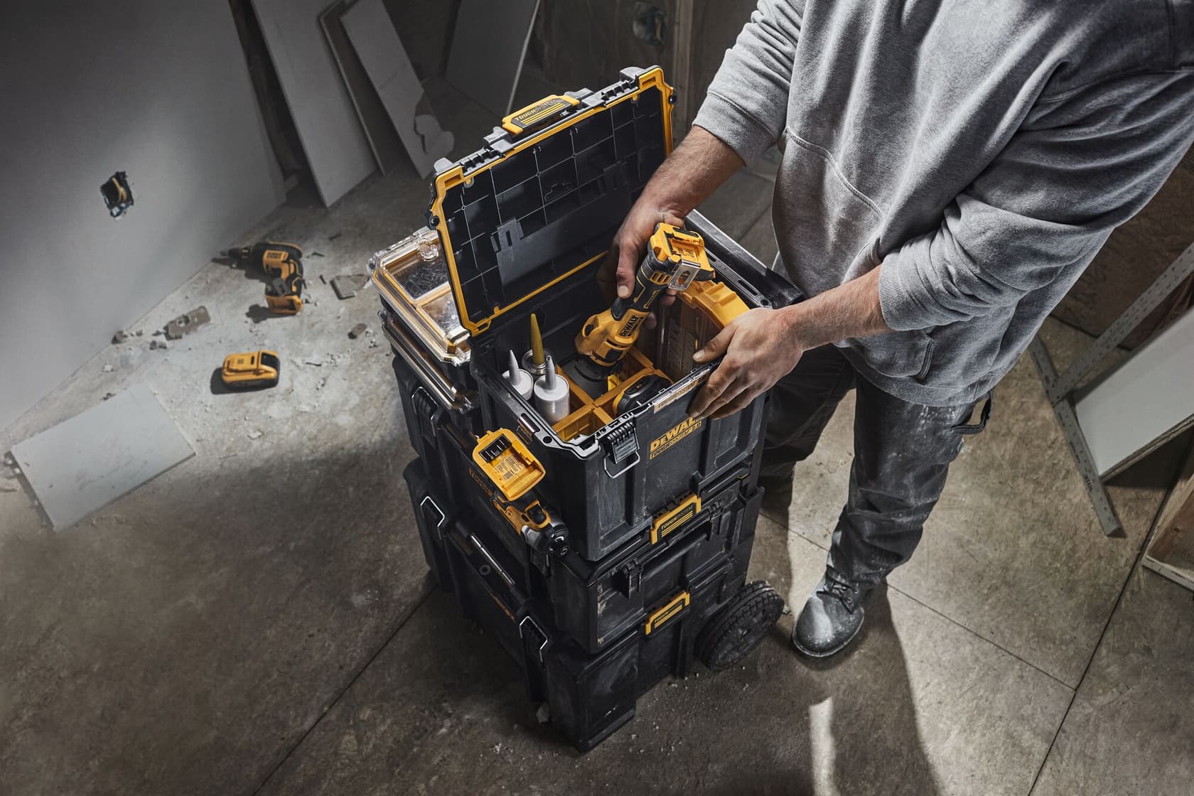 Una persona está de pie junto a una pila de cajas de almacenamiento DEWALT resistentes, organizando herramientas dentro de una de las cajas. Las cajas son negras con detalles amarillos y están ubicadas en una obra en construcción. Herramientas eléctricas y accesorios son visibles tanto dentro de la caja como en el suelo cercano.