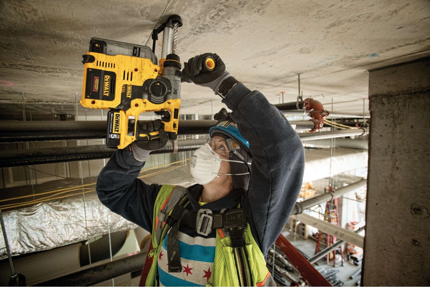 Un trabajador de la construcción con chaleco de seguridad, casco, guantes y mascarilla está utilizando un taladro percutor amarillo DEWALT para perforar el techo de concreto en un sitio de construcción.