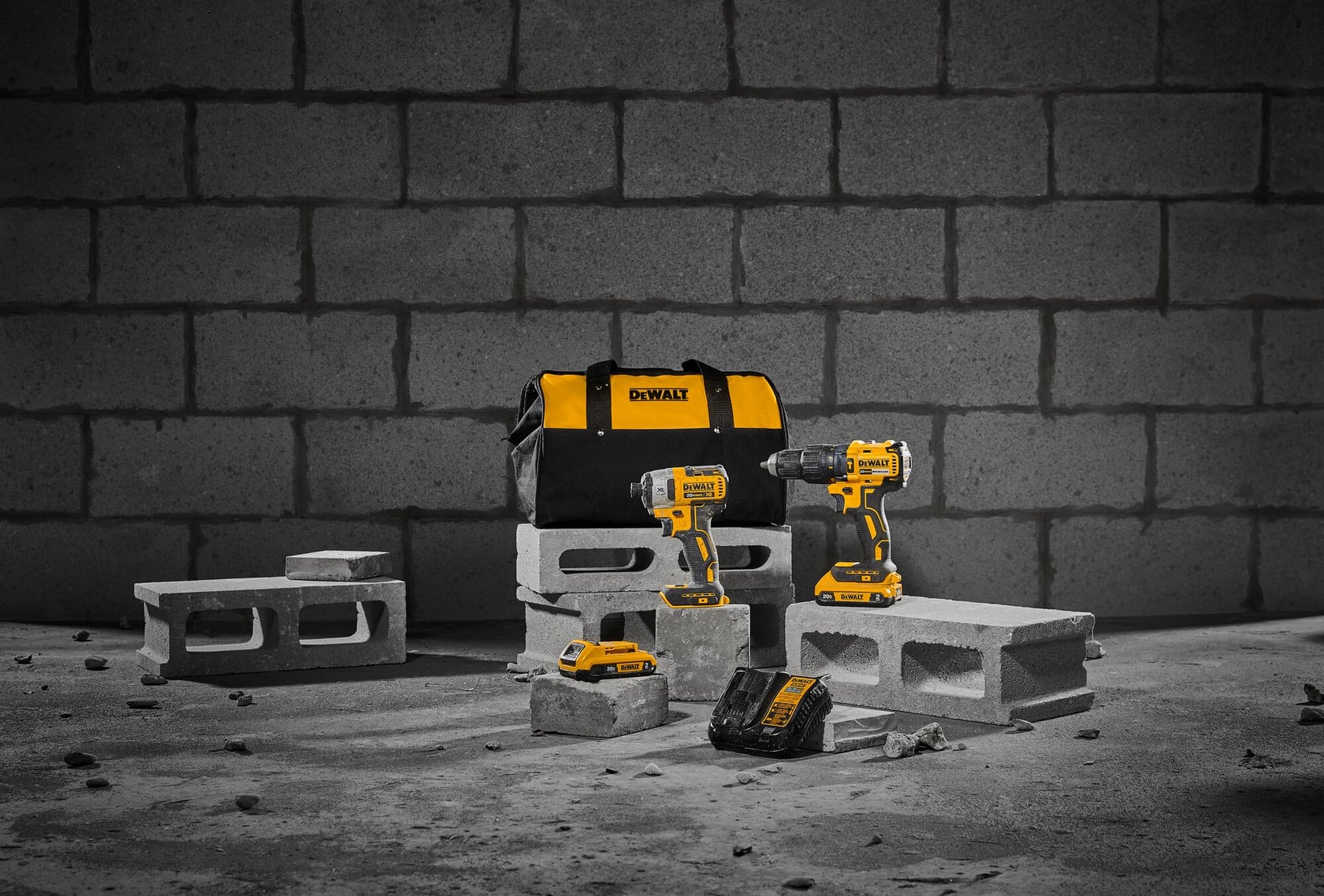 Un set de herramientas DEWALT que incluye una bolsa de transporte negra y amarilla, dos taladros inalámbricos, un paquete de batería y un cargador, exhibido sobre bloques de concreto frente a una pared de ladrillos.