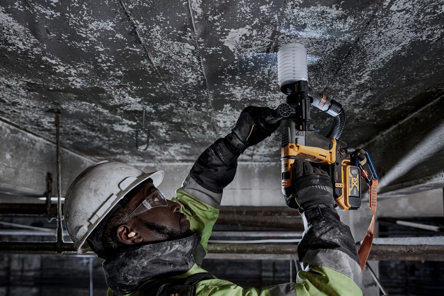 Un trabajador equipado con casco, guantes y gafas protectoras utiliza una herramienta eléctrica amarilla DEWALT para perforar un techo de concreto en un entorno industrial.