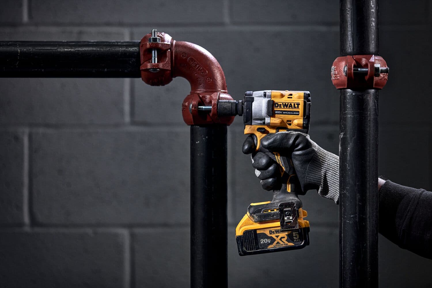 A person wearing black gloves is using a DEWALT cordless impact wrench to tighten or loosen a red pipe fitting on black industrial pipes in front of a grey concrete wall.