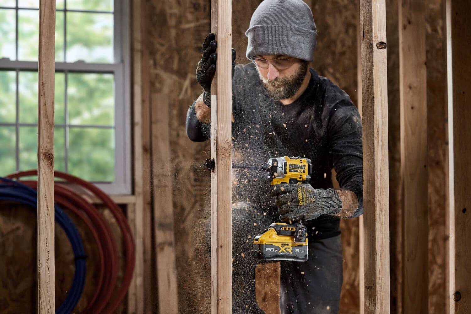 A person wearing gloves and a hat is using a DEWALT DCF860B cordless power tool to drill into a wooden frame in a construction setting. Sawdust is flying as the tool is operated.