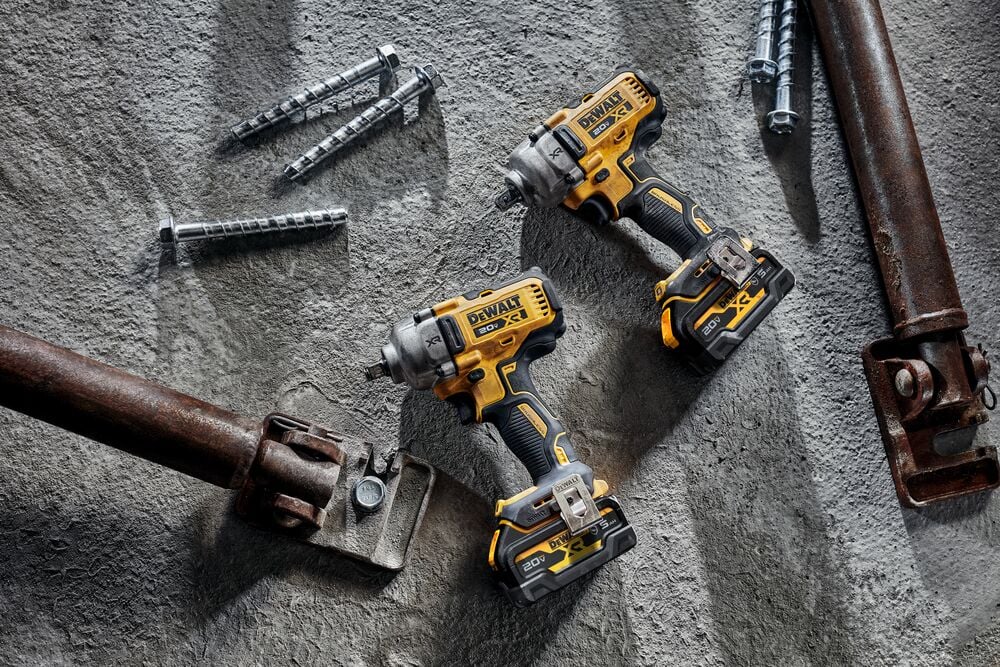 Dos llaves de impacto inalámbricas DEWALT con baterías de 20V están colocadas sobre una superficie de concreto junto a varios pernos metálicos grandes y componentes metálicos industriales.