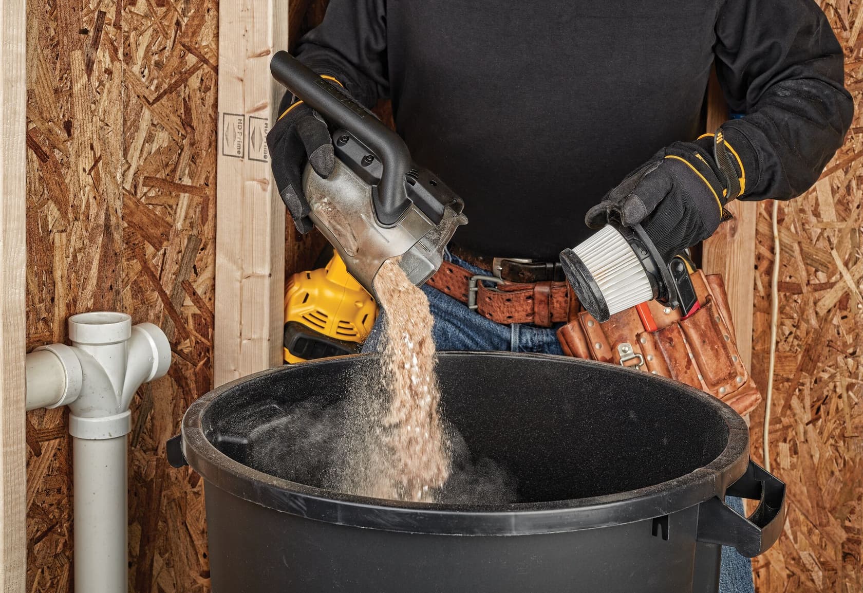 Una persona con guantes vacía polvo y residuos de un depósito de aspiradora DEWALT en un gran contenedor negro. También sostiene un filtro de aspiradora y lleva un cinturón de herramientas en la cintura. La escena tiene lugar en un taller con paredes de madera y tuberías visibles.